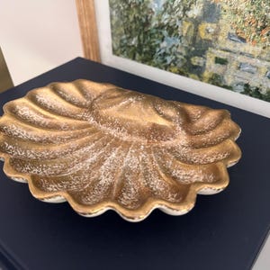 May include: A gold and white ceramic shell-shaped dish. The dish has a textured surface and a scalloped edge.