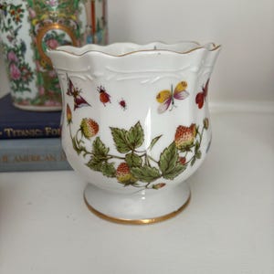 May include: White ceramic planter with a scalloped rim and gold trim. The planter is decorated with strawberries, green leaves, and colorful butterflies. The planter sits on a small pedestal base.