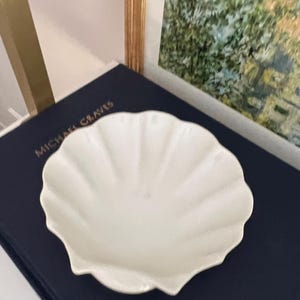 May include: A white ceramic bowl shaped like a seashell with a fluted edge. The bowl is sitting on a dark blue surface.