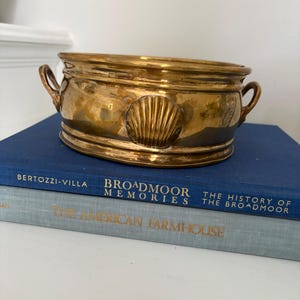 May include: A tarnished brass planter with a seashell design on the front, featuring two small handles. The planter sits atop two books with the titles "Broadmoor Memories" and "The American Farmhouse".