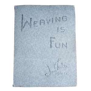 May include: A rectangular light blue woven textile featuring the dark blue stitched text "Weaving is Fun" and a signature that reads "Joy Tate 1946" in the lower right corner. The fabric has a consistent, textured, heathered appearance throughout.