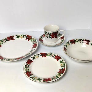 May include: A white ceramic Christmas dinnerware set featuring a red poinsettia and green holly design. The set includes an oval platter, a round plate, a bowl, a teacup, and a saucer, all with gold trim.