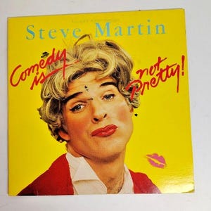 May include: A vintage album cover featuring Steve Martin in drag with bright red lipstick and a blonde wig. The yellow background has the text "Comedy is not Pretty!" in red and the title "Steve Martin" in blue.