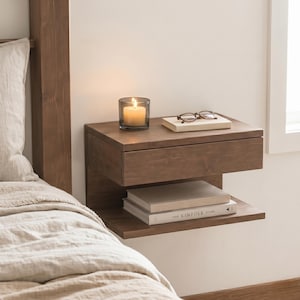 Puede incluir: Una mesita de noche flotante de madera con un cajón y una balda. Una vela encendida en un vaso, un libro y gafas descansan encima. Dos libros están en la balda de abajo. El marco de la cama y la ropa de cama son visibles.