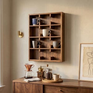 Puede incluir: Una estantería de pared de madera con múltiples compartimentos, que muestra varias tazas. Debajo se instala una estación de café, con un vertedor de vidrio, un molinillo de café, un tarro de frijoles y una taza en una bandeja de madera. Una obra de arte enmarcada está a la derecha.