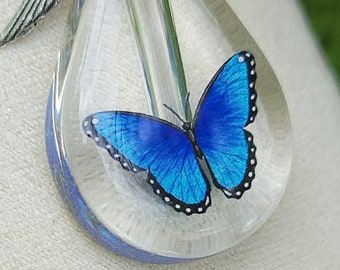 May include: A silver necklace with a clear glass pendant containing a blue butterfly. The pendant is shaped like a teardrop and has a silver angel wing charm attached to it.
