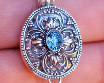 May include: An oval silver locket pendant featuring an intricate embossed floral pattern and a central oval blue glass cabochon. The locket has a beaded border and a small silver loop at the bottom, suspended from a thin silver chain necklace.