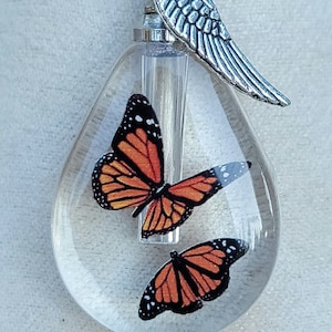 May include: A clear glass teardrop-shaped pendant with two orange and black butterfly images inside. The pendant is attached to a silver chain with a silver angel wing charm.