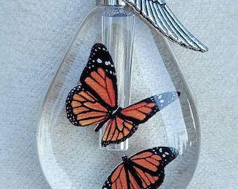 May include: A clear glass teardrop-shaped pendant with two orange and black butterfly images inside. The pendant is attached to a silver chain with a silver angel wing charm.