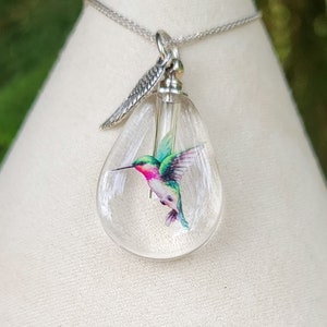 Puede incluir: Un collar de cadena de plata con un colgante de l&aacute;grima de vidrio transparente que presenta una ilustraci&oacute;n de colibr&iacute; colorida. El colgante tiene un dije de ala de plata unido a la parte superior.