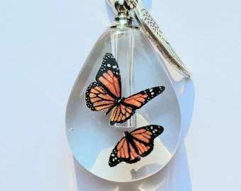 May include: A clear glass teardrop-shaped pendant with a silver chain and a silver wing charm. Inside the pendant are two orange and black butterfly images.