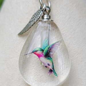 Puede incluir: Un collar con un colgante de cristal transparente en forma de l&aacute;grima, una cadena de plata y un dije de plata en forma de colibr&iacute;. El colibr&iacute; est&aacute; pintado en tonos de verde, rosa y azul.