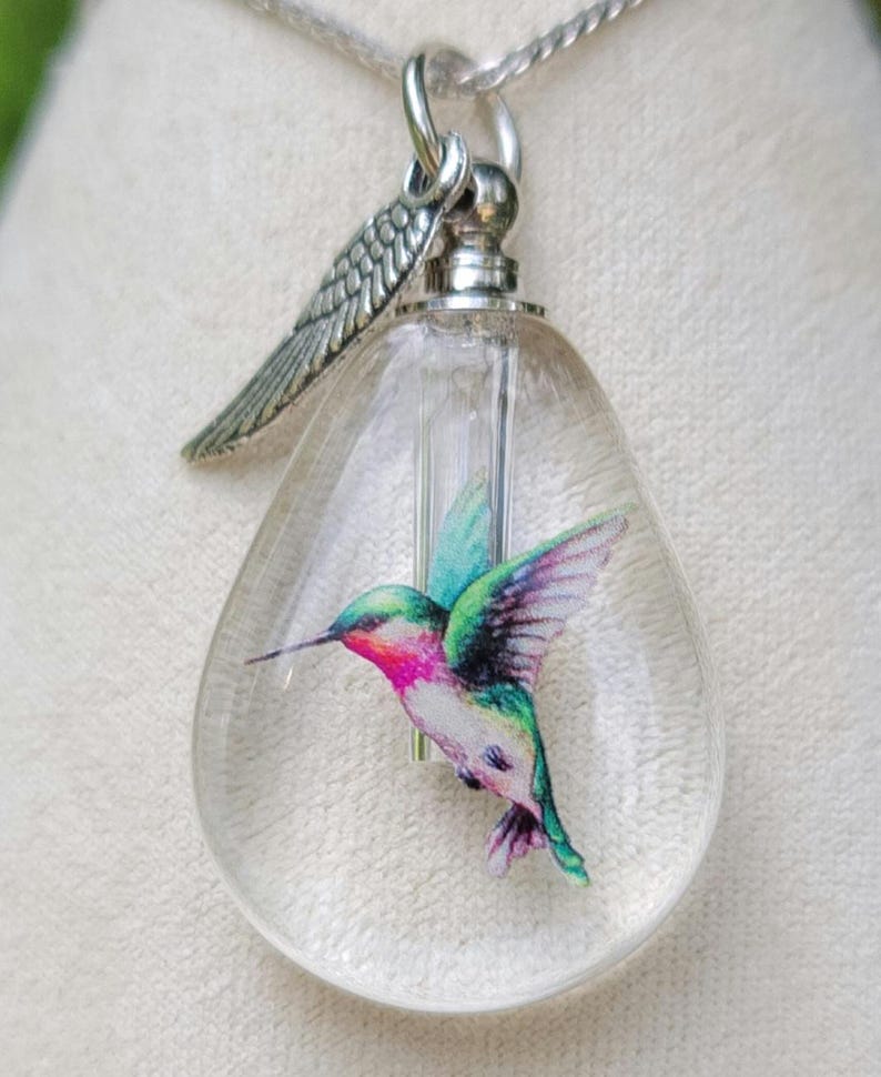 Puede incluir: Un collar con un colgante de cristal transparente en forma de l&aacute;grima, una cadena de plata y un dije de plata en forma de colibr&iacute;. El colibr&iacute; est&aacute; pintado en tonos de verde, rosa y azul.