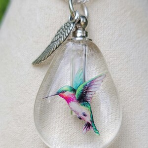 Puede incluir: Un collar con un colgante de vidrio transparente, una cadena de plata y un dije de ala de &aacute;ngel de plata. El colgante contiene una ilustraci&oacute;n colorida de colibr&iacute;.