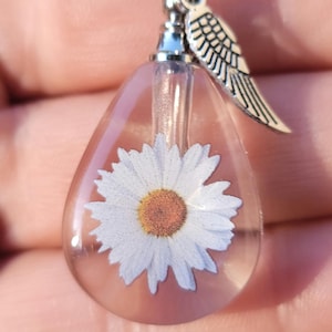 May include: A clear, teardrop-shaped pendant with a white daisy encased inside. The pendant has a silver-colored cap and a small wing charm. The daisy has a yellow center and white petals. The pendant is suspended from a silver chain.