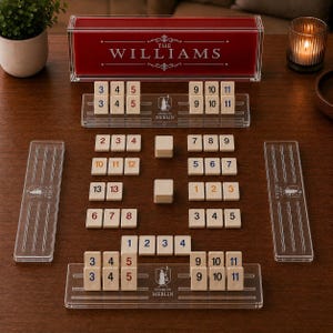 May include: A personalized red and clear acrylic game set with the name "THE WILLIAMS" printed on the lid. The game includes numbered tiles and acrylic tile holders. The game is set up on a dark brown wooden surface.
