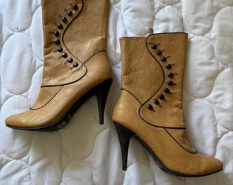 May include: A pair of tan leather ankle boots with black trim and heels. The boots have a button closure on the side and a pointed toe. The heels are approximately 7.6 cm tall.