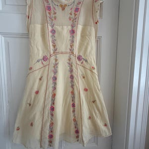 May include: Cream-colored sleeveless dress with floral embroidery in pink, purple, and orange. The dress features a high neckline and a flared skirt. The embroidery is concentrated along the bodice and down the front panels, creating a delicate and feminine look.