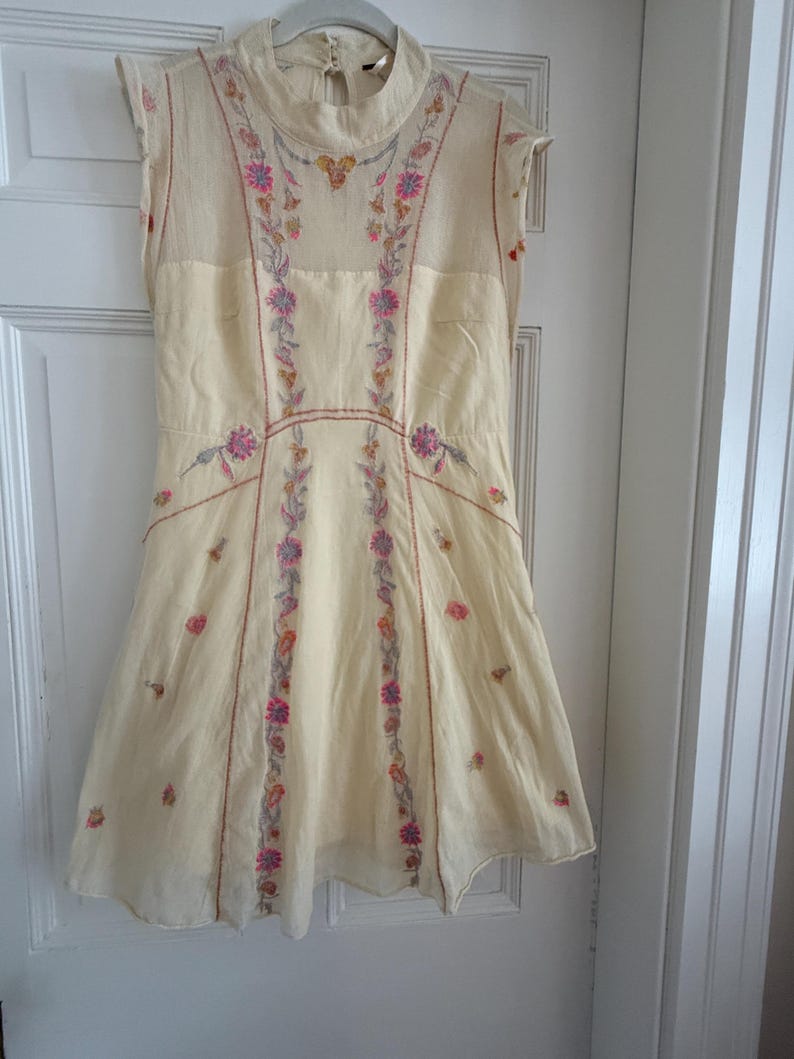 May include: Cream-colored sleeveless dress with floral embroidery in pink, purple, and orange. The dress features a high neckline and a flared skirt. The embroidery is concentrated along the bodice and down the front panels, creating a delicate and feminine look.