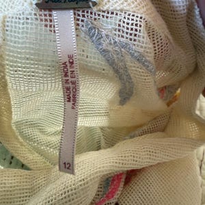May include: Close-up of a cream-colored, open-weave fabric with a Free People tag. A size 12 tag is visible. The fabric has a delicate, airy texture, with hints of pink and blue.