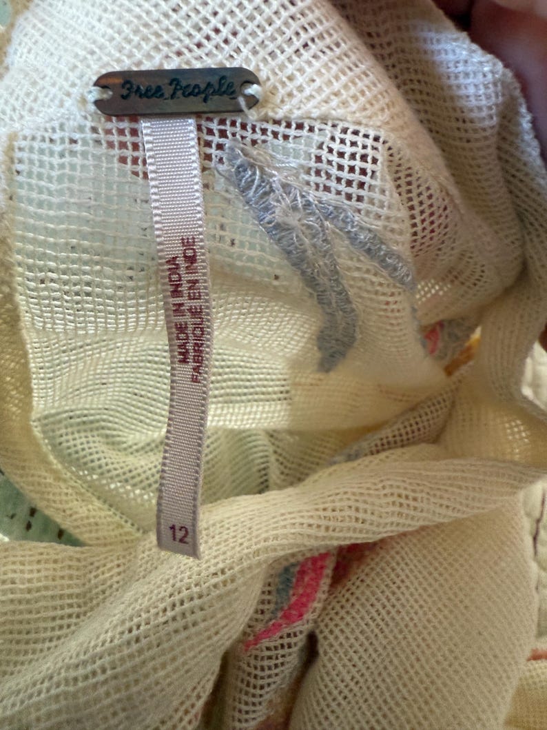 May include: Close-up of a cream-colored, open-weave fabric with a Free People tag. A size 12 tag is visible. The fabric has a delicate, airy texture, with hints of pink and blue.