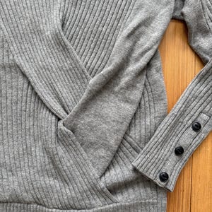 May include: A light gray ribbed knit sweater featuring a crossover front design and long sleeves. The sleeve cuffs are detailed with three small, round black buttons arranged in a vertical line along the outer edge of the ribbed fabric.