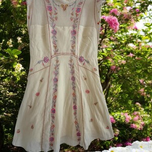 May include: Cream-colored sleeveless dress with floral embroidery in pink, purple, and green. The dress features a high neckline and a flared skirt. The embroidery is concentrated along the bodice and down the front, creating a delicate and feminine look.