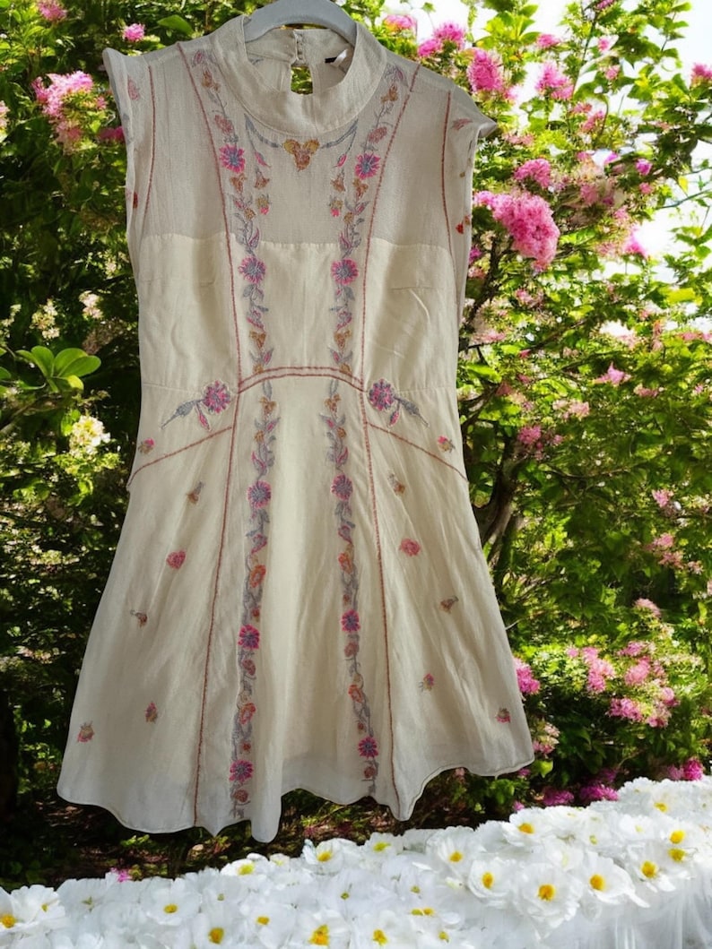 May include: Cream-colored sleeveless dress with floral embroidery in pink, purple, and green. The dress features a high neckline and a flared skirt. The embroidery is concentrated along the bodice and down the front, creating a delicate and feminine look.