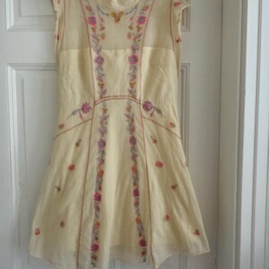 May include: A cream-colored sleeveless dress with a high neckline, adorned with floral embroidery in pink, purple, and yellow. The dress hangs on a hanger against a white door, showcasing its delicate design and flowing silhouette.