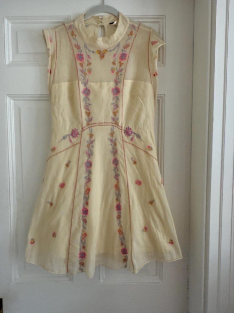 May include: A cream-colored sleeveless dress with a high neckline, adorned with floral embroidery in pink, purple, and yellow. The dress hangs on a hanger against a white door, showcasing its delicate design and flowing silhouette.