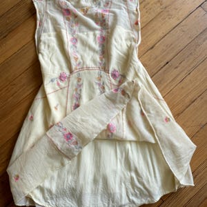 May include: A cream-colored sleeveless dress with floral embroidery in pink and yellow. The dress features a high neckline with a keyhole opening in the back and a flared skirt. The embroidery is concentrated on the bodice and along the seams.