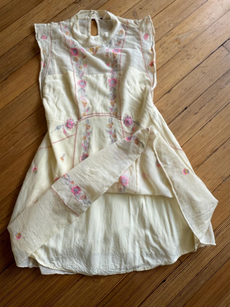 May include: A cream-colored sleeveless dress with floral embroidery in pink and yellow. The dress features a high neckline with a keyhole opening in the back and a flared skirt. The embroidery is concentrated on the bodice and along the seams.