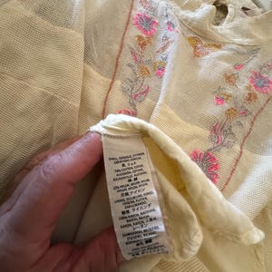 May include: Cream-colored, sheer top with floral embroidery in pink, yellow, and gray. The top features a high neckline and a delicate, open weave. The label shows the fabric composition.