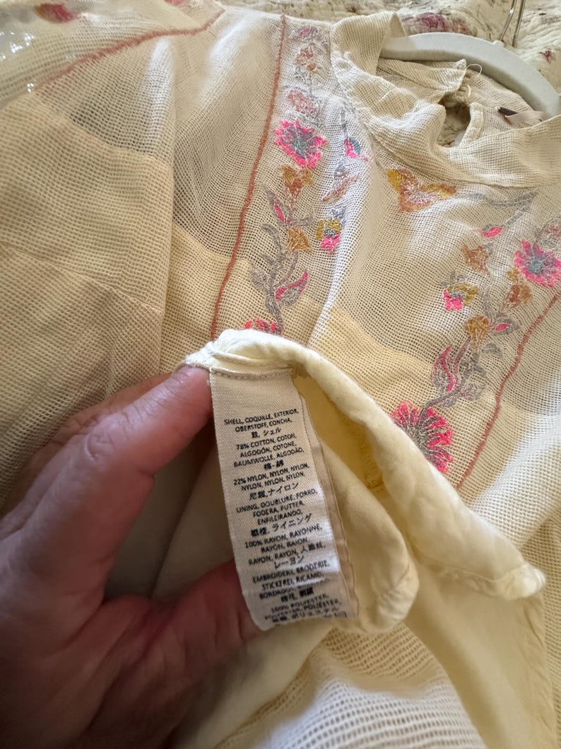 May include: Cream-colored, sheer top with floral embroidery in pink, yellow, and gray. The top features a high neckline and a delicate, open weave. The label shows the fabric composition.