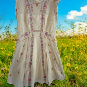 May include: Cream-colored sleeveless dress with floral embroidery in pink, purple, and green. The dress features a high neckline and a flared skirt. The embroidery is concentrated along the bodice and down the front panels, creating a delicate, feminine look.