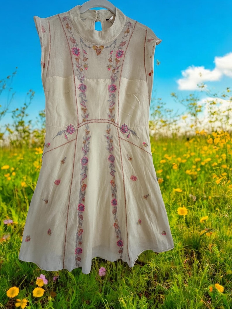 May include: Cream-colored sleeveless dress with floral embroidery in pink, purple, and green. The dress features a high neckline and a flared skirt. The embroidery is concentrated along the bodice and down the front panels, creating a delicate, feminine look.