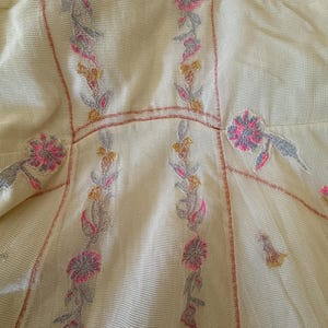 May include: Close-up of a cream-colored garment with floral embroidery. The embroidery features pink, gray, and yellow flowers with green leaves. The garment has a mesh-like texture and a fitted design.