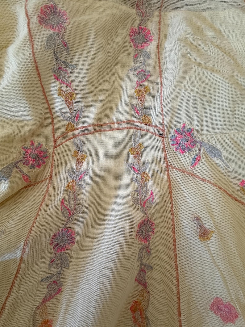 May include: Close-up of a cream-colored garment with floral embroidery. The embroidery features pink, gray, and yellow flowers with green leaves. The garment has a mesh-like texture and a fitted design.