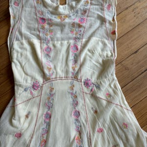 May include: Cream-colored dress with floral embroidery in pink, gray, and yellow. The sleeveless dress features a high neckline and a keyhole back. The embroidery is concentrated down the front and around the waist, with delicate floral accents.
