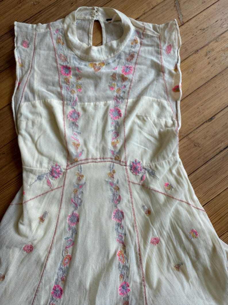 May include: Cream-colored dress with floral embroidery in pink, gray, and yellow. The sleeveless dress features a high neckline and a keyhole back. The embroidery is concentrated down the front and around the waist, with delicate floral accents.