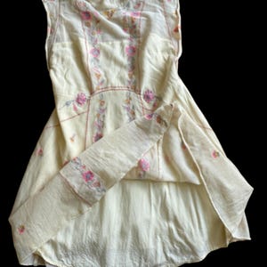 May include: A cream-colored vintage dress with floral embroidery in pink and yellow. The dress features a high neckline, keyhole back, and cap sleeves. The embroidery is concentrated on the bodice and along the seams, creating a delicate and feminine look.
