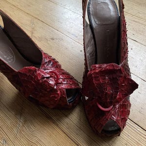 May include: A pair of red leather heeled shoes with a slingback strap. The shoes are made of a textured leather that resembles snakeskin. The shoes have a pointed toe and a high heel.