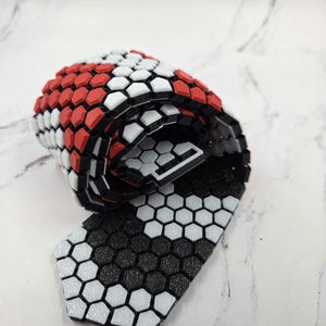 Pode incluir: Gravata enrolada com um padrão geométrico de formas hexagonais vermelhas, brancas e pretas. A gravata é feita de um material flexível e tem uma superfície texturizada. O design é moderno e atraente.
