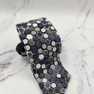 Pode incluir: Uma gravata estampada com um design geométrico. A gravata apresenta um padrão repetitivo de pequenas formas hexagonais em tons de roxo, cinza, branco e preto. A gravata é presa com uma faixa elástica preta.