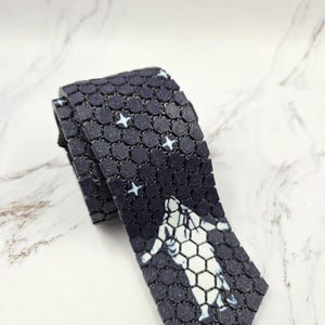 Pode incluir: Uma gravata azul marinho com um padrão hexagonal repetido e detalhes de estrelas brancas. A gravata apresenta um design de astronauta branco com um padrão hexagonal. A gravata está dobrada e repousa sobre uma superfície de mármore branco e cinza.