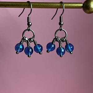 May include: A pair of dangle earrings featuring three blue glass beads on each earring. The beads are attached to a silver-colored metal ring and hook. The earrings are displayed against a pink background.