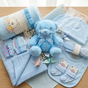 May include: A baby gift set in various shades of blue, featuring a teddy bear, blanket, hat, bib, socks, and a brush and comb set. The items are arranged on light blue paper, creating a cohesive and charming presentation.