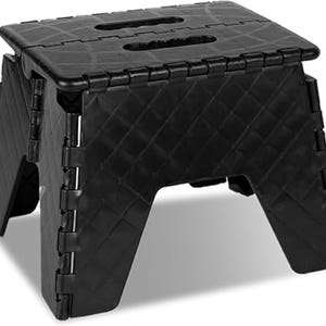 May include: A black, folding step stool. The stool is made of plastic and has a diamond pattern on the sides and top. It features two handle cutouts on the top surface and folds flat for storage. The stool is shown against a white background.
