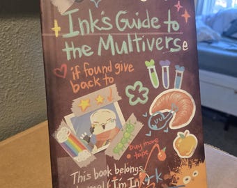 May include: A hardback book titled Ink's Guide to the Multiverse with a dark purple cover featuring colourful stickers, hand-drawn illustrations of a croissant, an apple, test tubes, and text that reads This book belongs to me! I'm Ink.