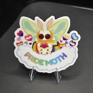 May include: A die-cut sticker featuring a fluffy white moth with large brown eyes and pale green wings. The moth wears a rainbow-striped cape and holds a small red heart. Surrounding the moth are various pride flag-colored hearts and the text PRIDE MOTH.